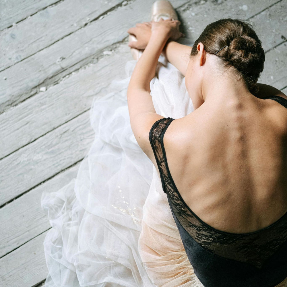 A ballet dancer stretching on a wooden floor, wearing a black lace leotard and a white tutu. Her hair is styled in a bun as she leans forward, elegantly showcasing her skin, back muscles, spine, and ribs.