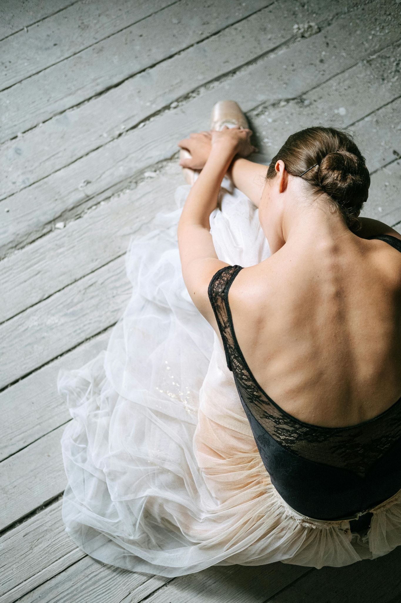 A ballet dancer stretching on a wooden floor, wearing a black lace leotard and a white tutu. Her hair is styled in a bun as she leans forward, elegantly showcasing her skin, back muscles, spine, and ribs.
